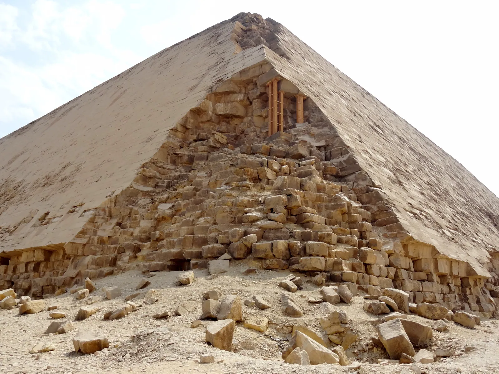 Ett hörn av Knäckta pyramiden: slät beklädnad upptill, bortplockade fyllnads- och kärnstenar nedtill och träställningen upp till norringången. Utmaning: kontrollera vid ett hörn att beklädnadsskiften lutar inåt (6–9° längst ned) i stället för att ligga vågrätt.
