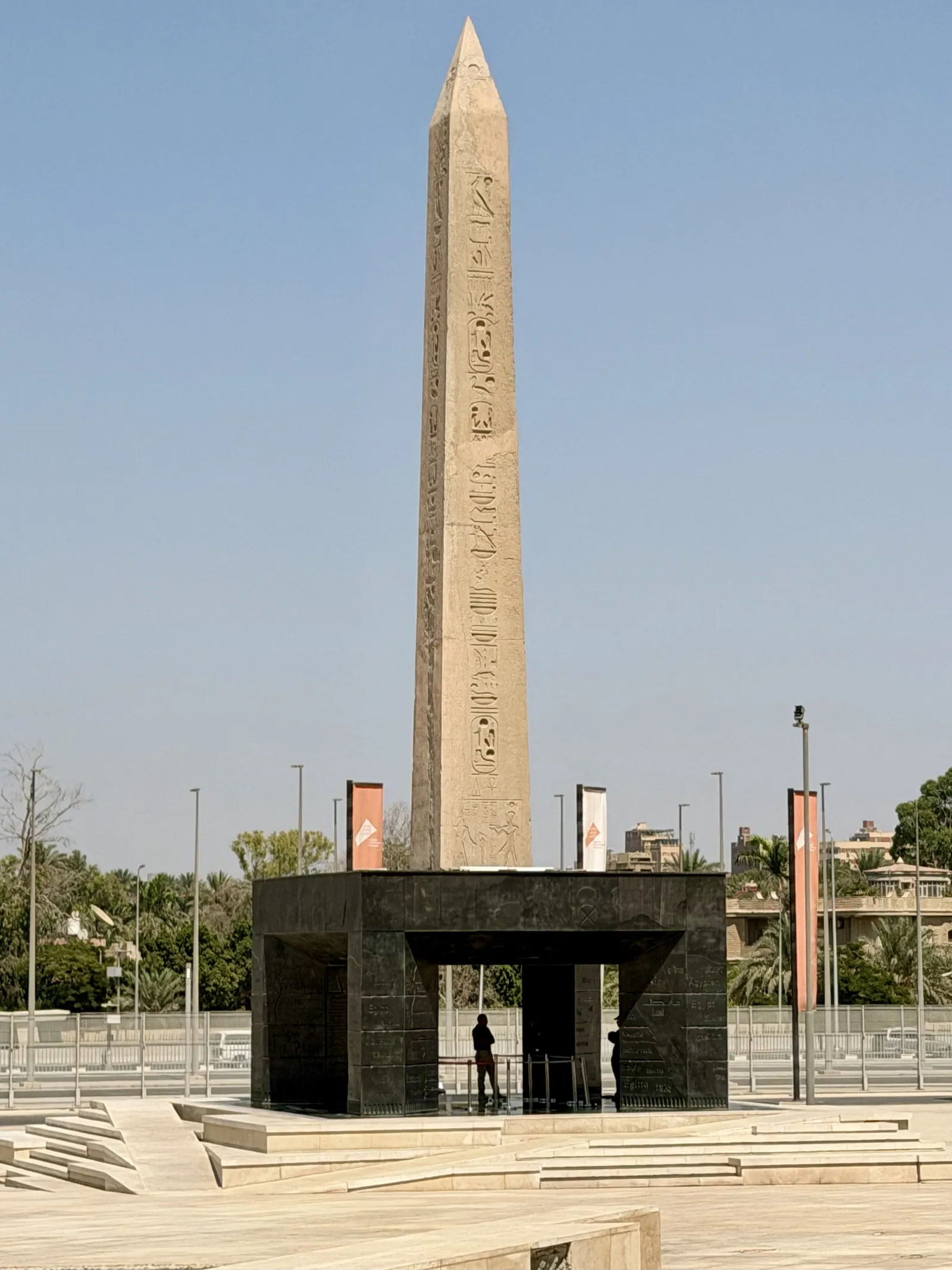 Den hängande obelisken på GEM:s torg (foto aug. 2026): Ramses II:s 16 meter höga och 87 ton tunga granitobelisk från Tanis, upplyft på en stålram så att man kan se kartuscherna på undersidan.