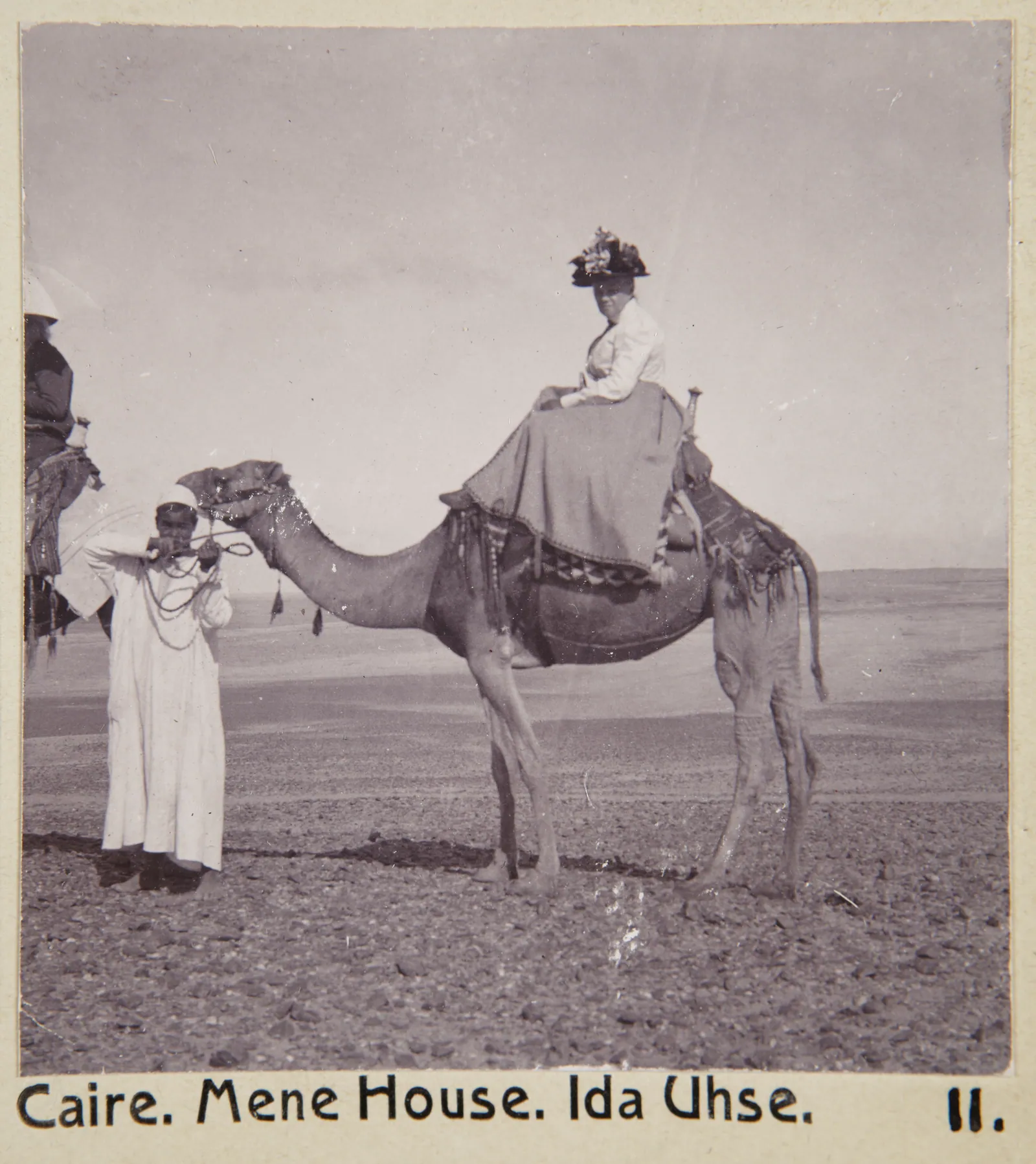 ”Mena House. Ida Uhse.” – ur albumet från den stockholmska familjen von Hallwyls resa genom Egypten och Sudan (5 nov 1900–29 mars 1901): en dam i bredbrättad hatt på kamel i öknen vid hotellet Mena House nedanför platån.